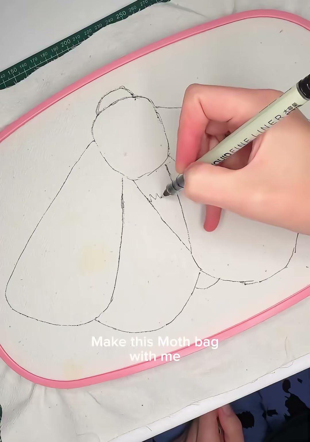 Step 01 — A hand drawing the outline of a large moth onto fabric stretched in a pink embroidery hoop, with the words "Make this Moth bag with me" visible on screen