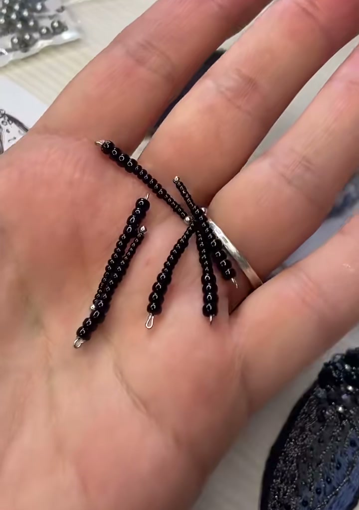Step 05 — Six identical beaded legs resting in the palm of a hand, each made of black seed beads strung on wire, ending in tiny silver jump rings