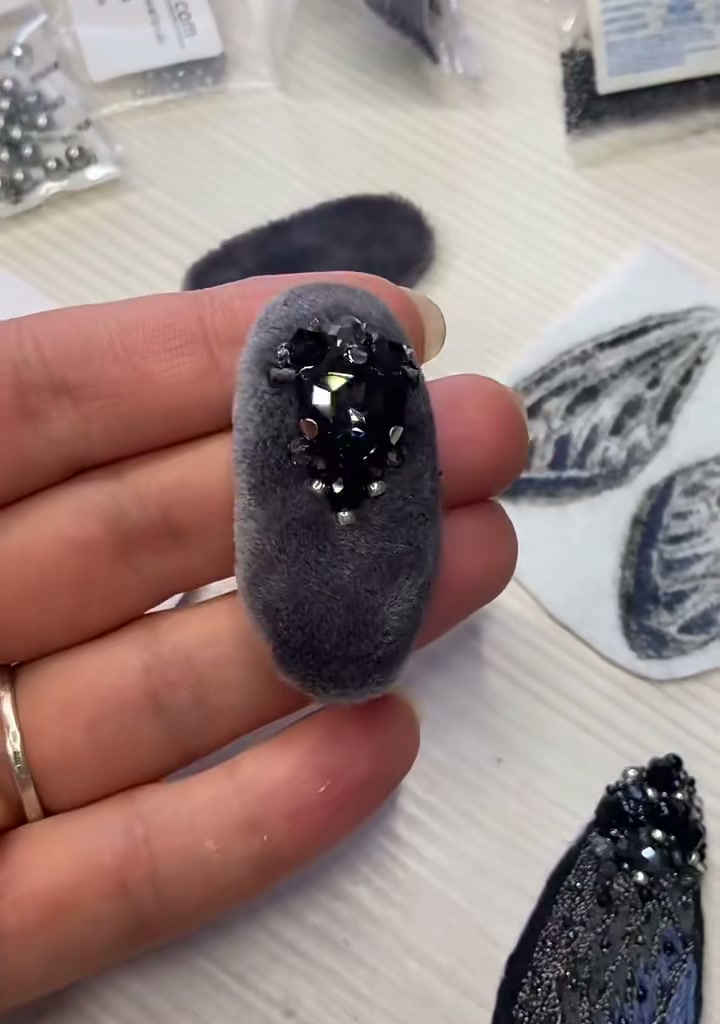 Step 06 — The completed cicada brooch resting on a hand, shown from a slight side angle: black encrusted body, midnight-blue wings fanning out, two silver antennae pointing up, six beaded legs draping forward