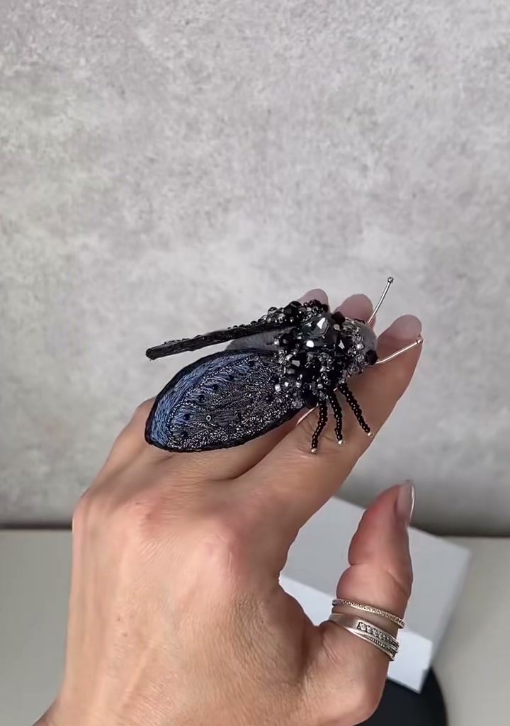 Step 07 — The completed cicada brooch held between fingers, showing the full spread of midnight-blue filigree wings, the towering rhinestone thorax, and the six articulated legs reaching forward