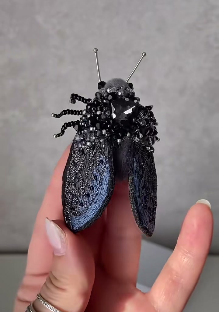 Step 08 — The brooch tilted upward, revealing the underside: the soft gray felt belly, the silver pin back, and the six beaded legs splaying outward like spokes