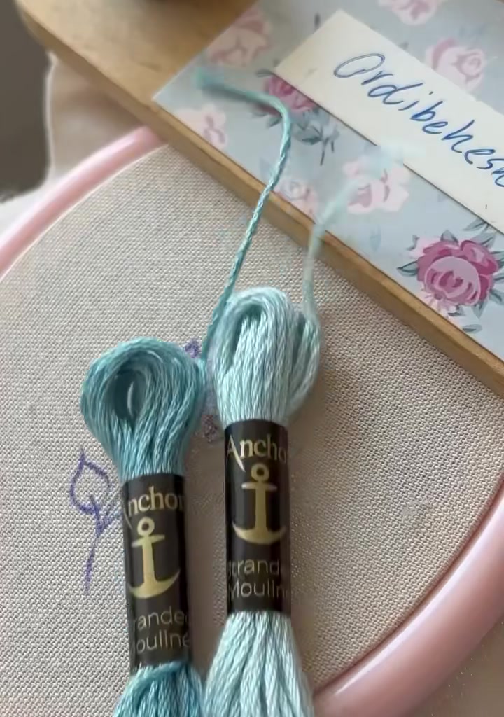 Two skeins of Anchor embroidery floss in blue shades placed on fabric inside a pink hoop