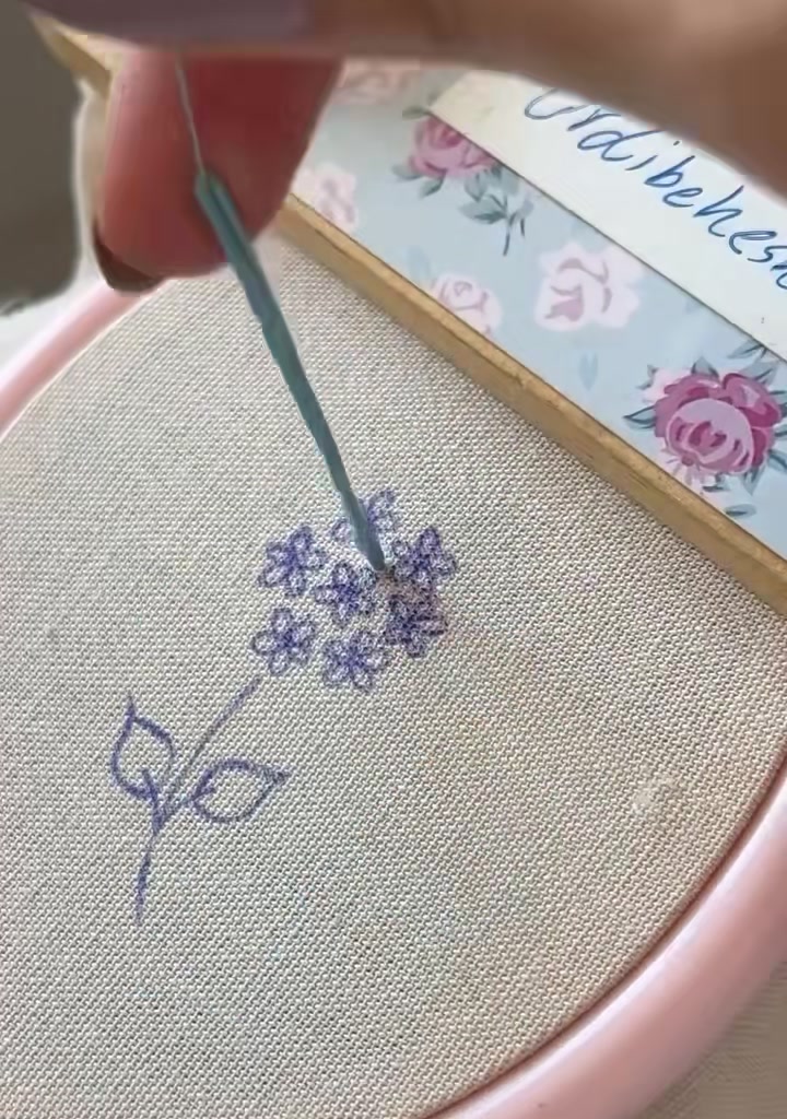 A hand bringing the needle up through the fabric at the center of the drawn flower